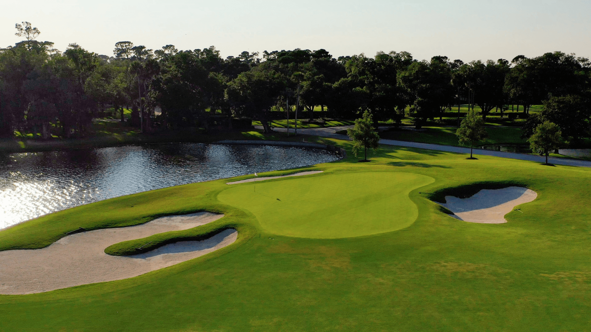 Lakeside green at Plantation Golf Course with calm water beside a well-manicured fairway, trees and shrubs lining the edge under a clear sky