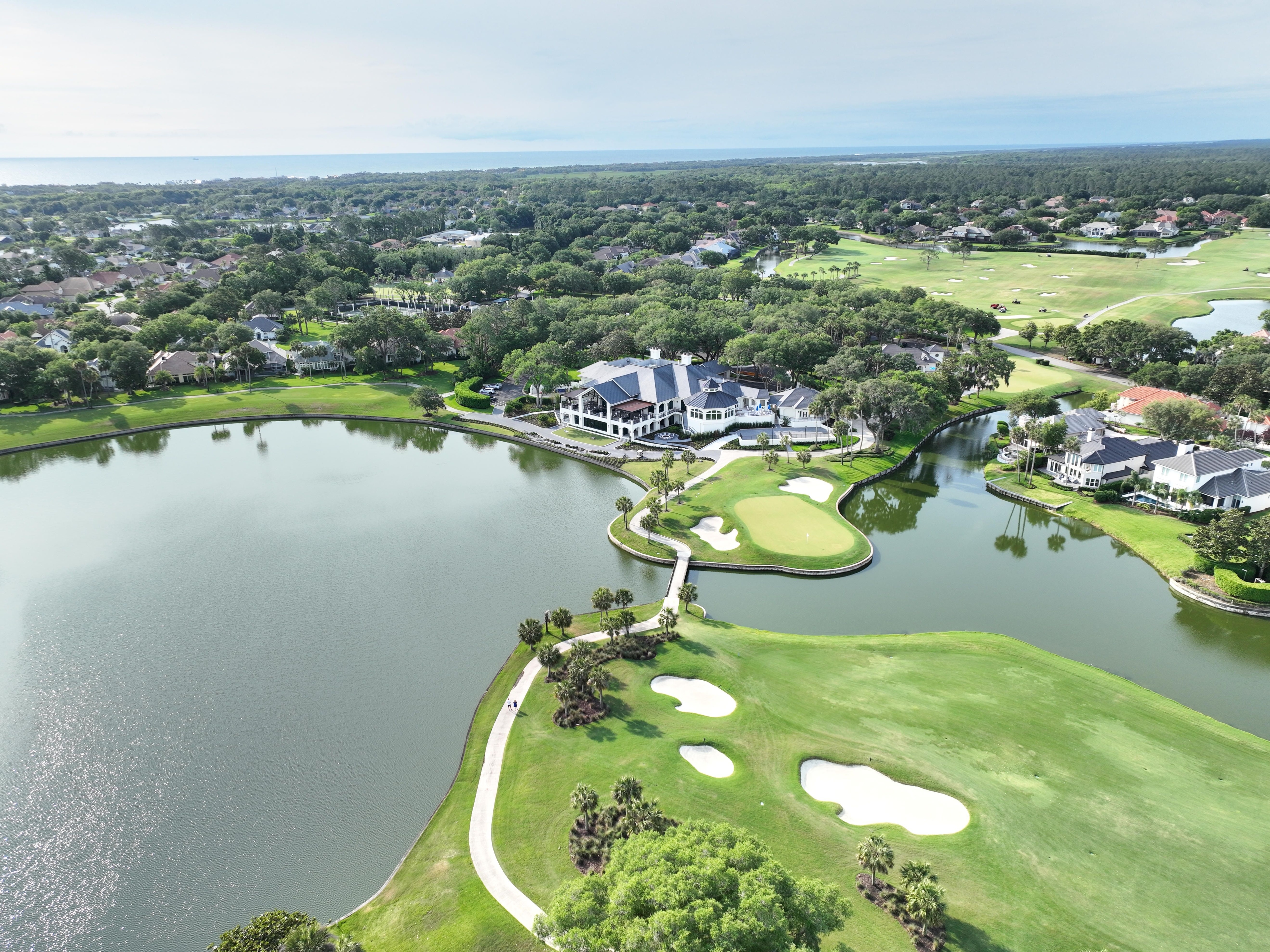 Aerial view of a lush green plantation-style golf course with fairways, greens, sand bunkers, and surrounding trees under clear skies