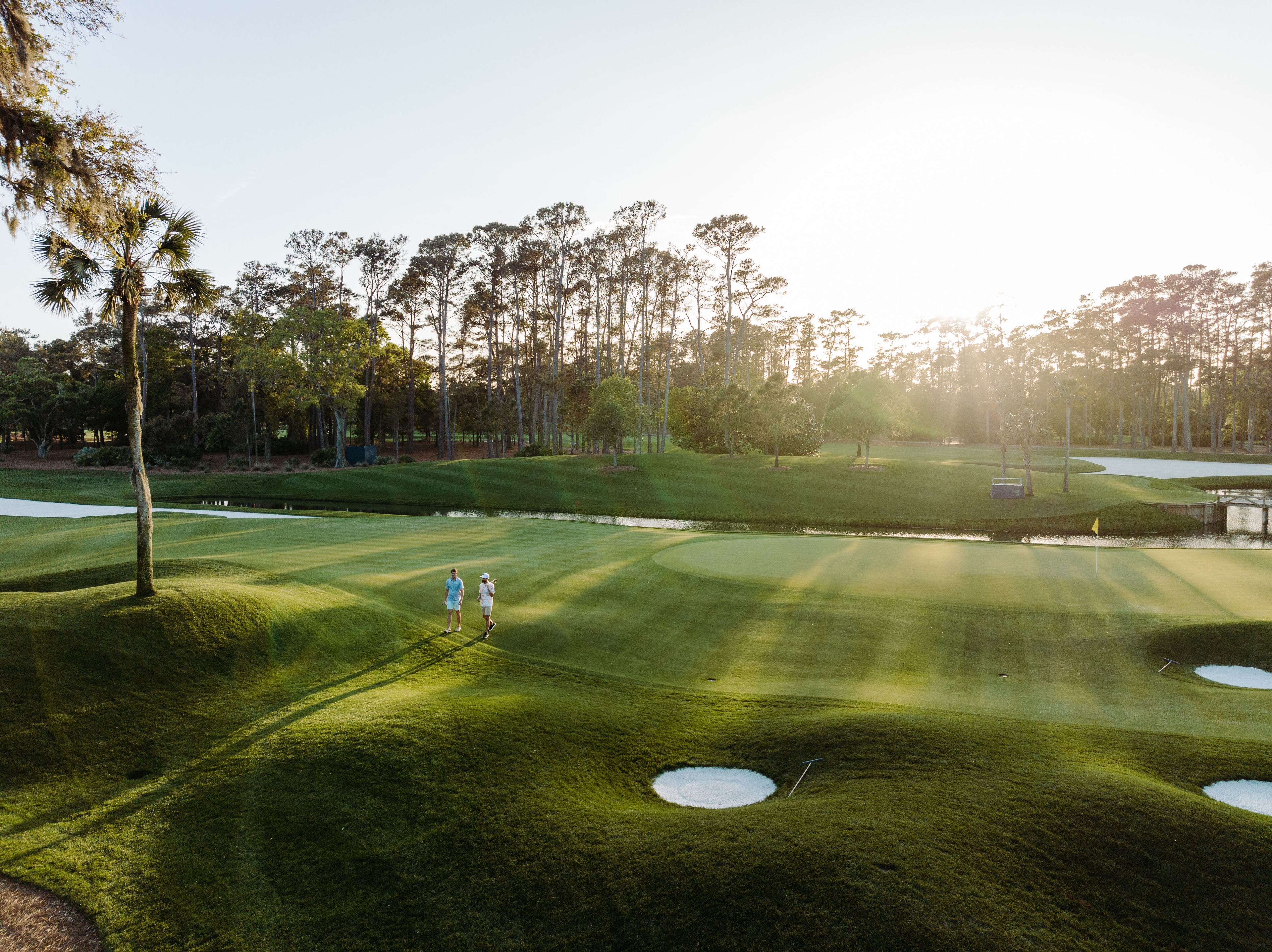 Sunlit sawgrass fairway with neatly mowed grass, scattered trees, and long shadows under a clear sky on a bright, calm day
