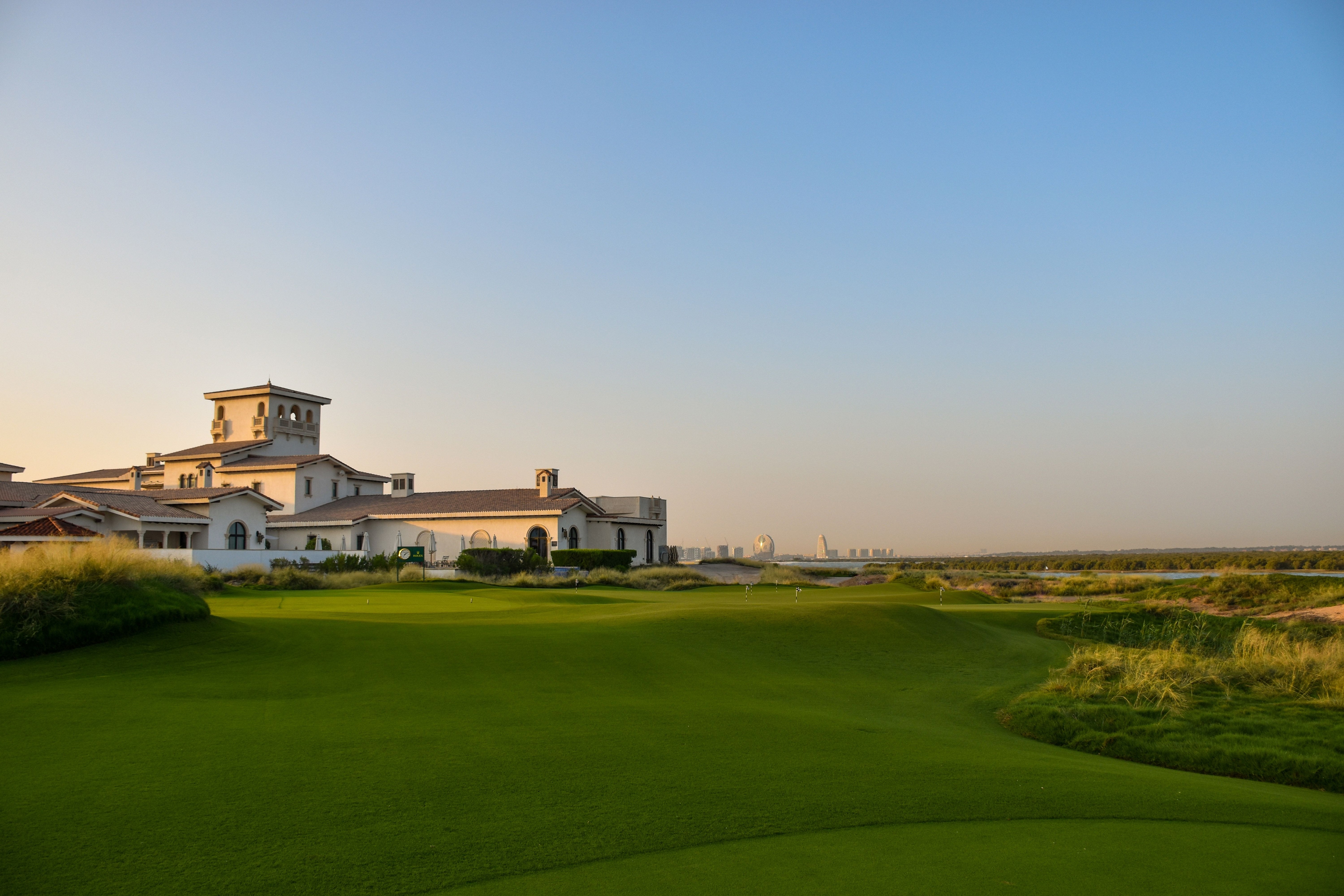 Aerial view of Yas Links golf course in Dubai with lush green fairways, sand bunkers, and water features beside the coastline under clear skies